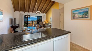 A kitchen with a black granite countertop, white cabinets, and a dishwasher. There is a dining area with a table and chairs, and a view of the outdoors through a large window. - Maui Kamaole #L-203 Full Ocean View, A/C Throughout, Wifi, Great Location (Kihei)