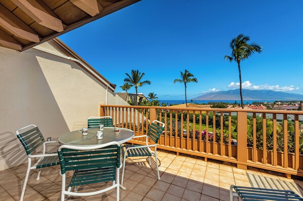 A balcony with a table and chairs, a view of palm trees and mountains, and a wooden railing.