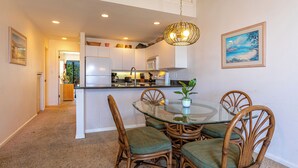A dining area with a glass table and wicker chairs, a kitchen in the background, and a painting on the wall. - Maui Kamaole #L-203 Full Ocean View, A/C Throughout, Wifi, Great Location (Kihei)