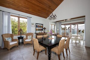 A dining area with a dark wooden table and wicker chairs, a side table with a lamp, and a view of the ocean through large windows.