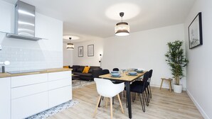 Una cocina moderna con armarios blancos, encimera de madera y estufa integrada. Hay una zona de comedor con mesa de madera, sillas negras y una planta en maceta.