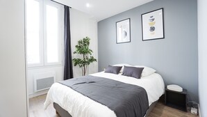 Un dormitorio moderno con una cama, una mesita de noche pequeña, una planta y dos obras de arte en la pared.