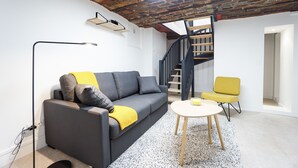 Una sala de estar moderna con un sofá gris, cojines amarillos, una mesita redonda y un sillón amarillo. En el fondo se ve una escalera con barandilla de madera.