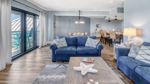 A modern living room with a blue sofa, a wooden coffee table with starfish and wooden plates, and a dining area in the background. - Gulf view, three bedroom, two bath condo in Mexico Beach. (Mexico Beach)