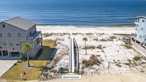 A beach entrance with a wooden boardwalk leading to the beach, a palm tree, and a building with a grey facade. - Gulf view, three bedroom, two bath condo in Mexico Beach. (Mexico Beach)
