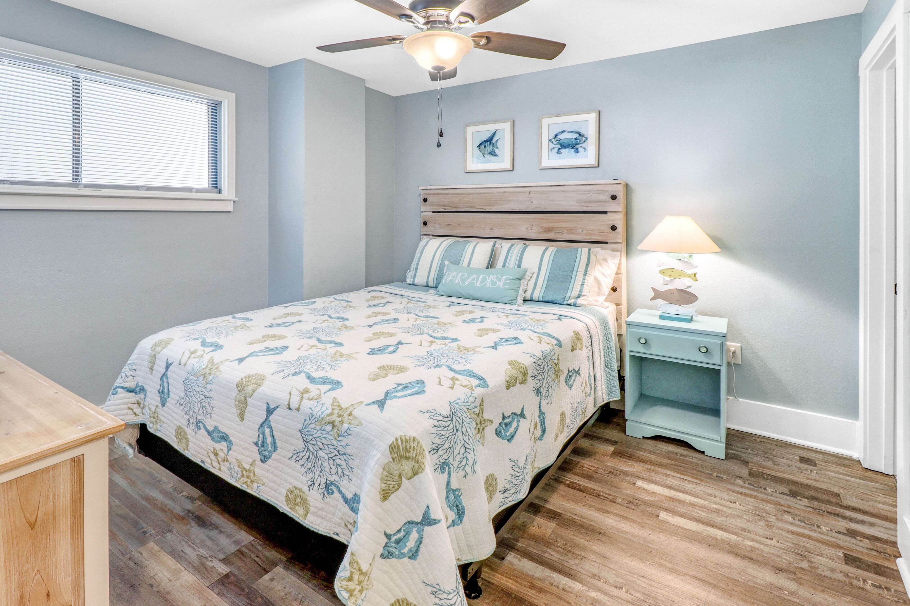 A bedroom with a bed, wooden headboard, a ceiling fan, a nightstand with a lamp, and a window with blinds.