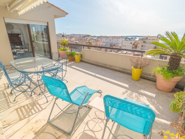 Een balkon met een glazen tafel en blauwe stoelen, plantenbakken en uitzicht op een stadsbeeld.