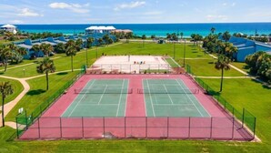 Tenniskenttä, jonka ympärillä on vehreää kasvillisuutta ja jolta avautuu esteetön näkymä merelle.