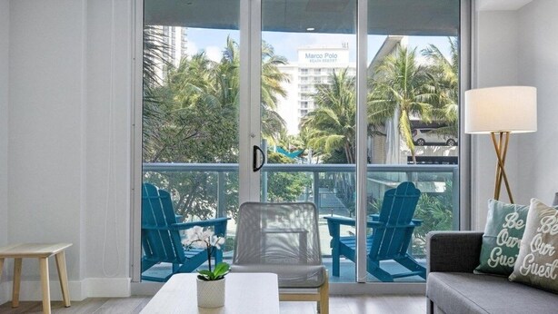 A balcony with a view of palm trees and a building labeled "Marco Polo Beach Resort." There are two blue chairs and a table with a plant. - 2br Luxury Ocean Reserve With Park Views! 2 Bedroom Condo by RedAwning (Sunny Isles Beach)