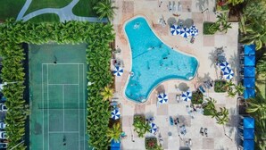 An aerial view of a resort with a large irregularly shaped swimming pool surrounded by lounge areas with umbrellas, a tennis court, and palm trees. - 2br Luxury Ocean Reserve With Park Views! 2 Bedroom Condo by RedAwning (Sunny Isles Beach)