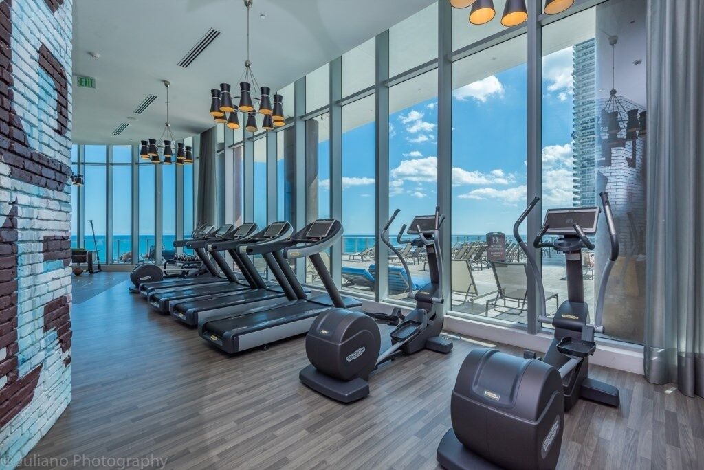 Un gimnasio con cintas de correr, elípticas y equipo de pesas, amplias ventanas que ofrecen vistas del cielo y un edificio.
