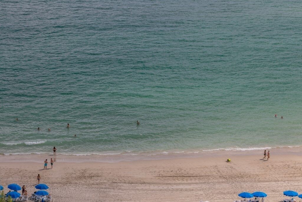 Uma praia com pessoas nadando e caminhando, e vários guarda-sóis azuis na areia.