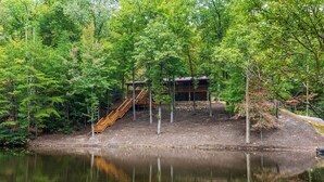 A cabin with a wooden deck and stairs leading to a deck area, surrounded by dense forest and a calm lake. - Beside Still Water 2 - 2 Bedrooms, 2 Baths, Sleeps 6 Home by RedAwning (Cosby)