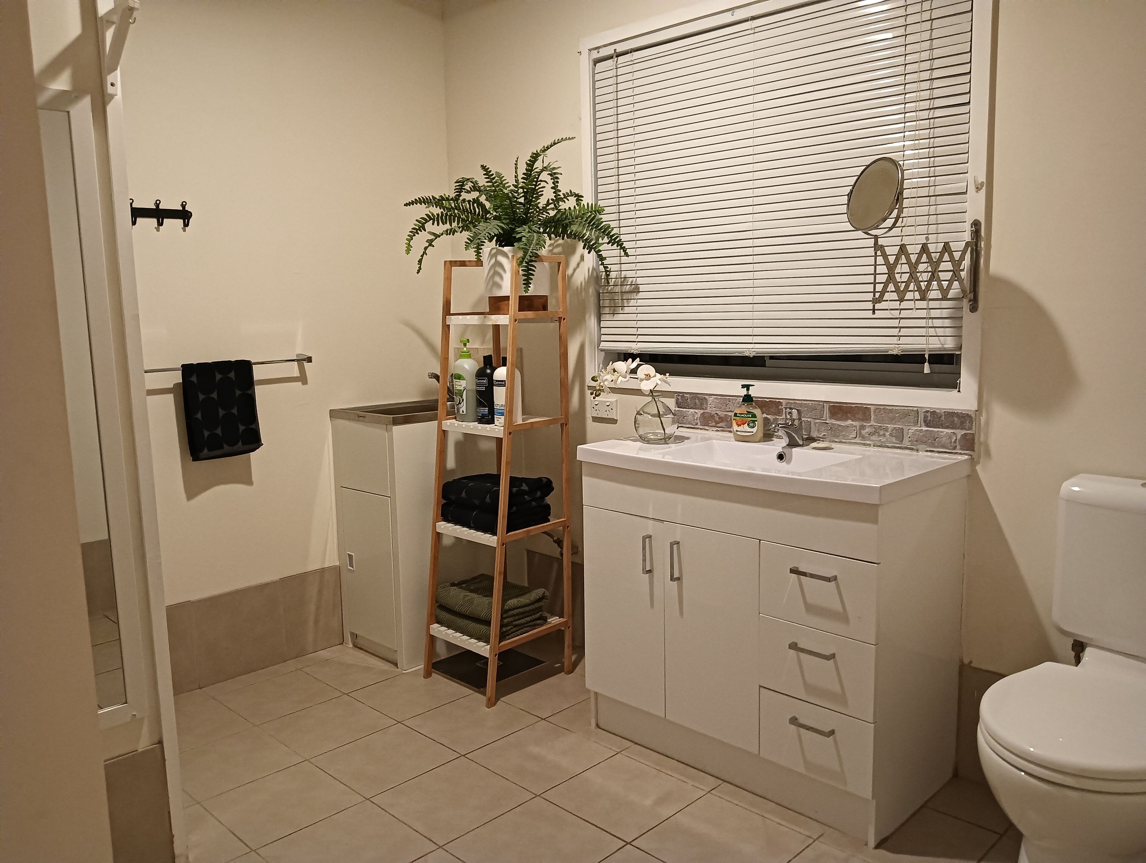 Une salle de bain avec un lavabo blanc, un miroir, des toilettes, un porte-serviettes et une étagère en bois où sont rangés les articles de toilette.