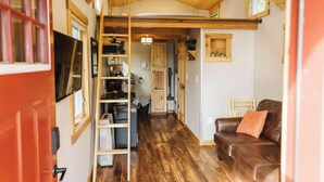 Ein gemütlicher, kompakter Wohnraum mit Holzboden, einem braunen Ledersofa, einem an der Wand montierten Fernseher und einer Küchenzeile mit Schränken und Spüle.