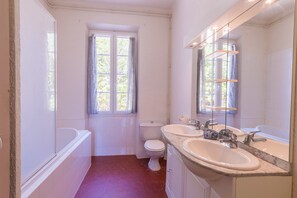 Un baño con bañera, dos lavamanos, inodoro y ventana con cortinas.