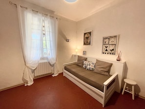 Ein kleines, gemütliches Schlafzimmer mit einem Einzelbett, einem Fenster mit durchsichtigen Vorhängen, einem Heizkörper und Wandbildern.