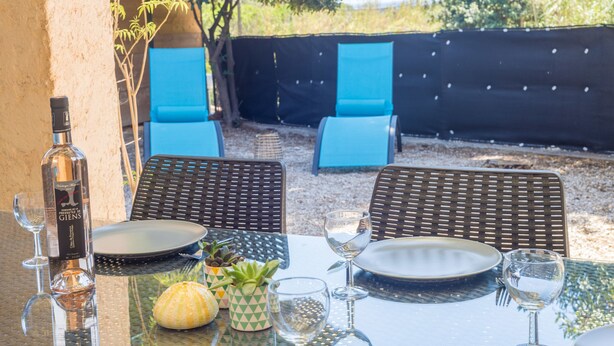 An outdoor dining area with a table set for two, including plates, glasses, and a bottle. There are two blue lounge chairs and a small potted plant on the table. - Le Mas, Provencal house between sea and vineyards (Hyères)