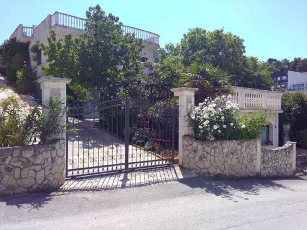 A gated entrance with stone walls and a metal gate, surrounded by lush greenery and a white building in the background.