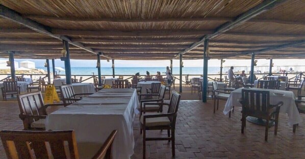 An outdoor dining area with tables and chairs, a thatched roof, and a view of the beach.