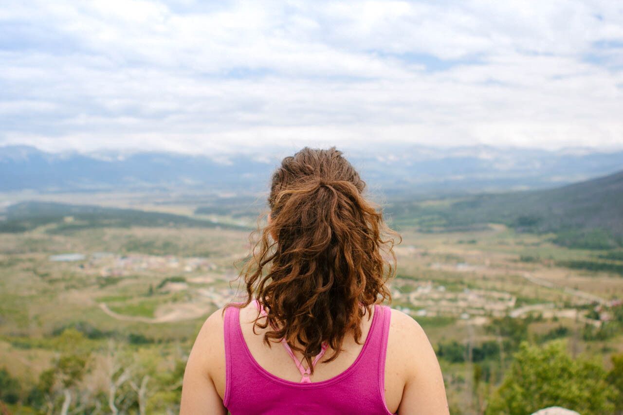 A person with curly hair from behind, wearing a pink tank top, overlooking a valley with scattered buildings and greenery.
