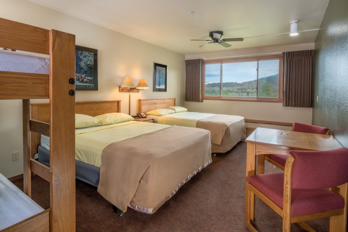 A hotel room with two beds, a table, a chair, a ceiling fan, and a window with a view.