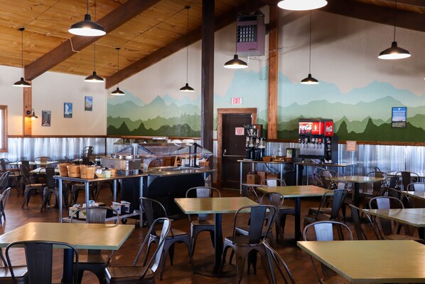 Un restaurante con altos techos de madera, mesas y sillas de metal, y un mural de montañas en la pared.