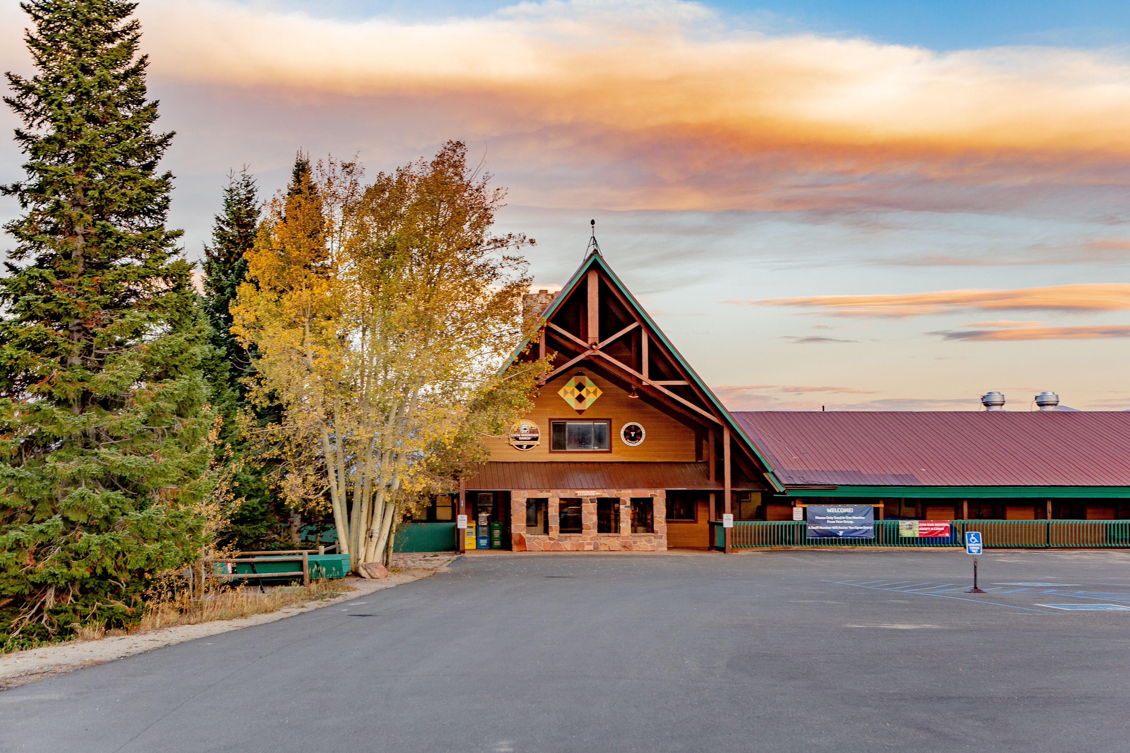 A lodge with a red roof and a stone facade, surrounded by trees and a parking lot.