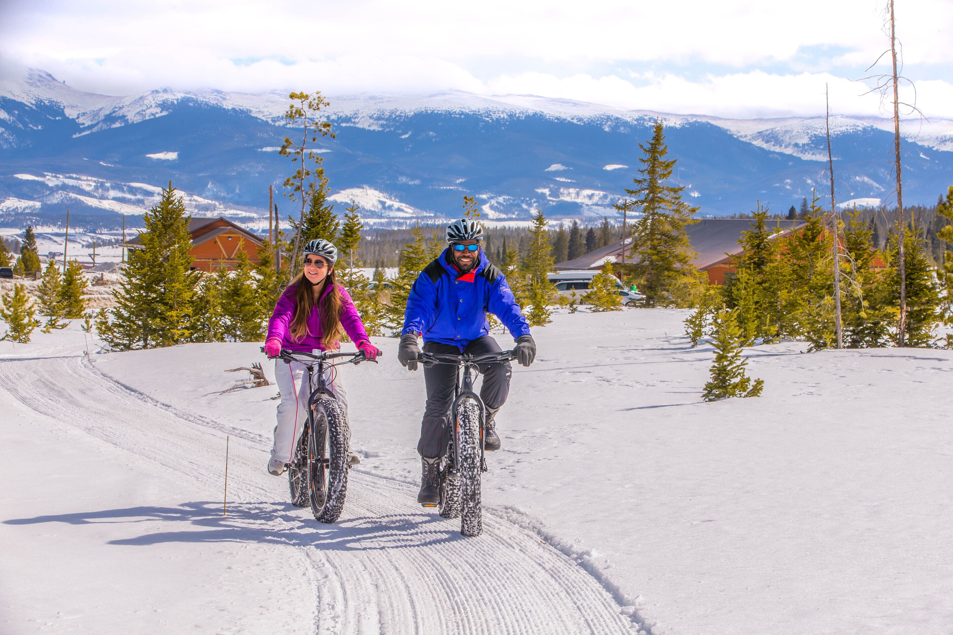Deux personnes à vélo dans un paysage enneigé, avec des montagnes en arrière-plan.