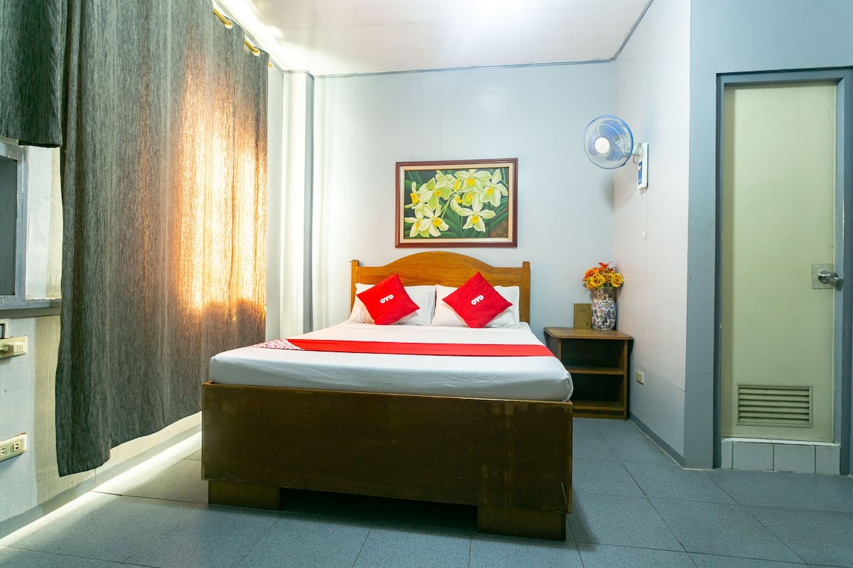 A hotel room with a wooden bed, a small bedside table with a vase, a wall-mounted fan, and a door.