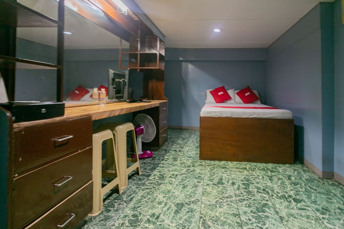 A compact hotel room with a bed, a desk with a computer, a chair, a small stool, and a fan.