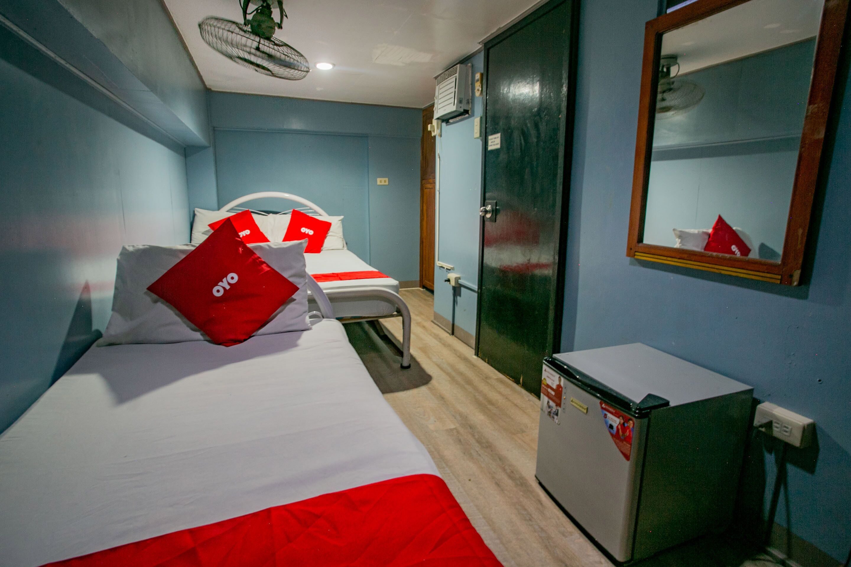 A room with two beds, a small refrigerator, a mirror, and a ceiling fan.
