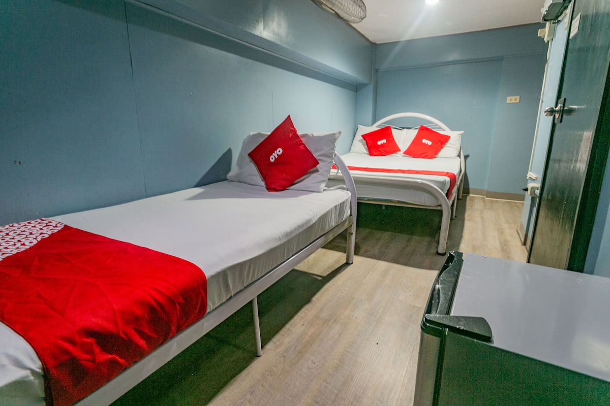 A small, compact room with two single beds, each with a red pillow and a white blanket. There is a small table and a door to the right.