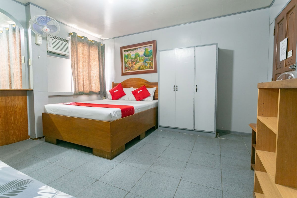 A room with a wooden bed, white curtains, a ceiling fan, a wardrobe, and a painting on the wall.
