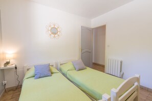 Um quarto com duas camas de solteiro, uma mesa de cabeceira com abajur, um adorno na parede e um radiador.
