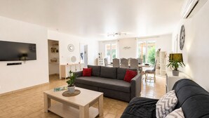 Ein modernes Wohnzimmer mit Flachbildfernseher, grauem Sofa mit roten Kissen, Couchtisch aus Holz und einem Essbereich mit Korbstühlen.
