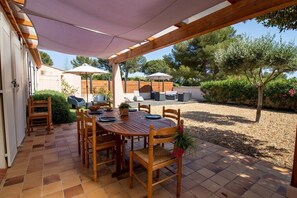 A covered patio with a wooden table and chairs, surrounded by a garden with trees and shrubs.