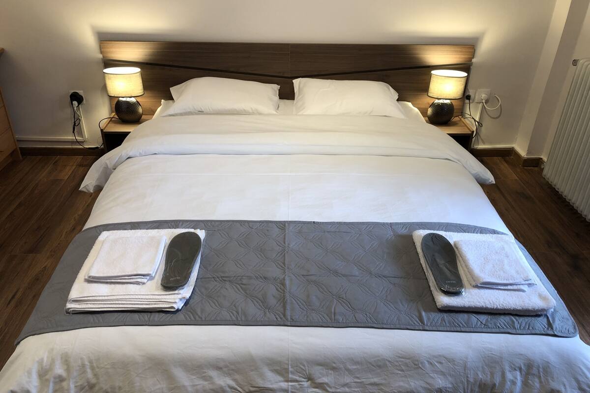 A neatly made bed with white linens, two bedside lamps, and a grey quilted bedspread.