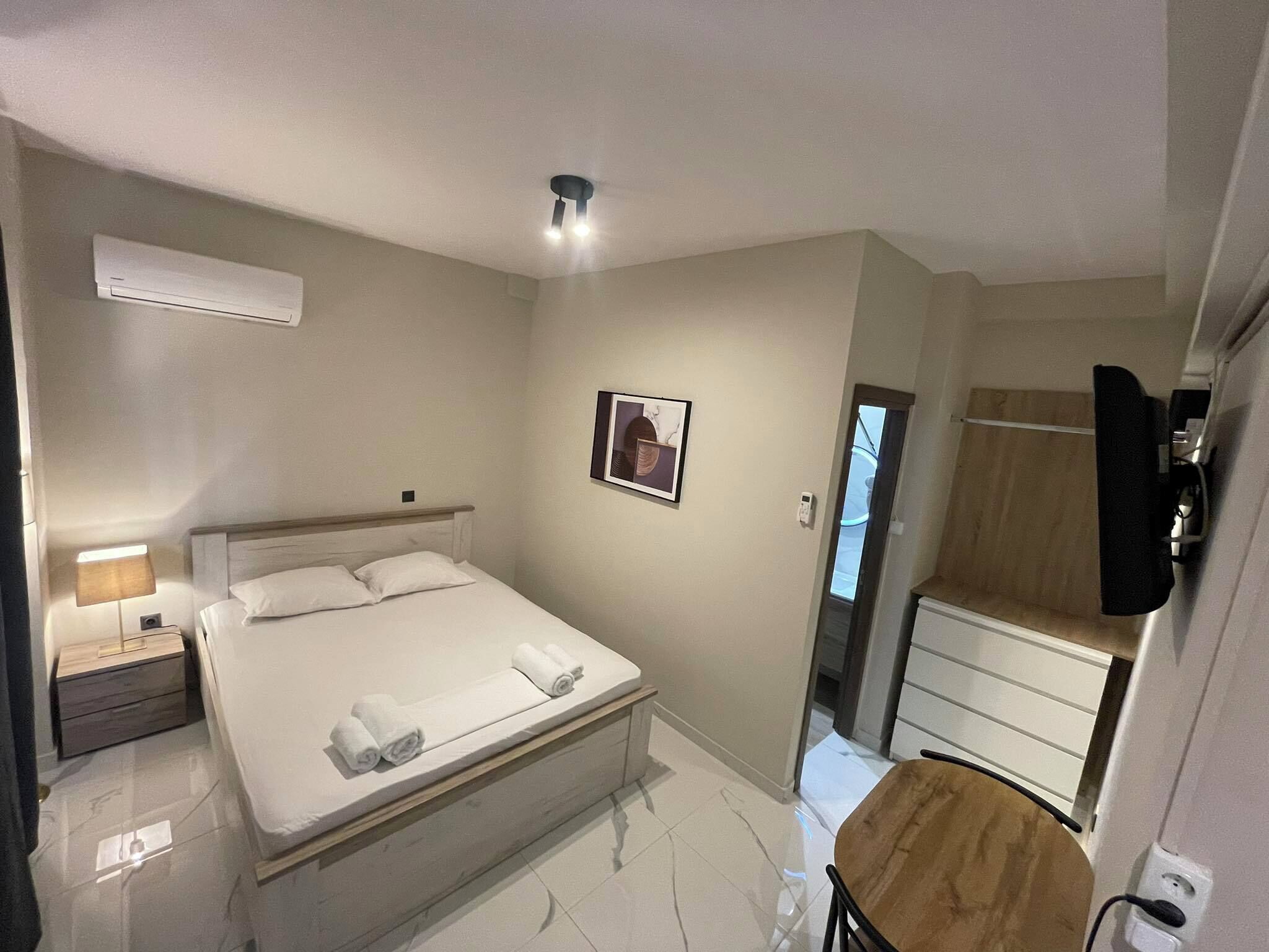 A hotel room with a bed, bedside table, lamp, air conditioning unit, framed picture, and a small table with a chair.
