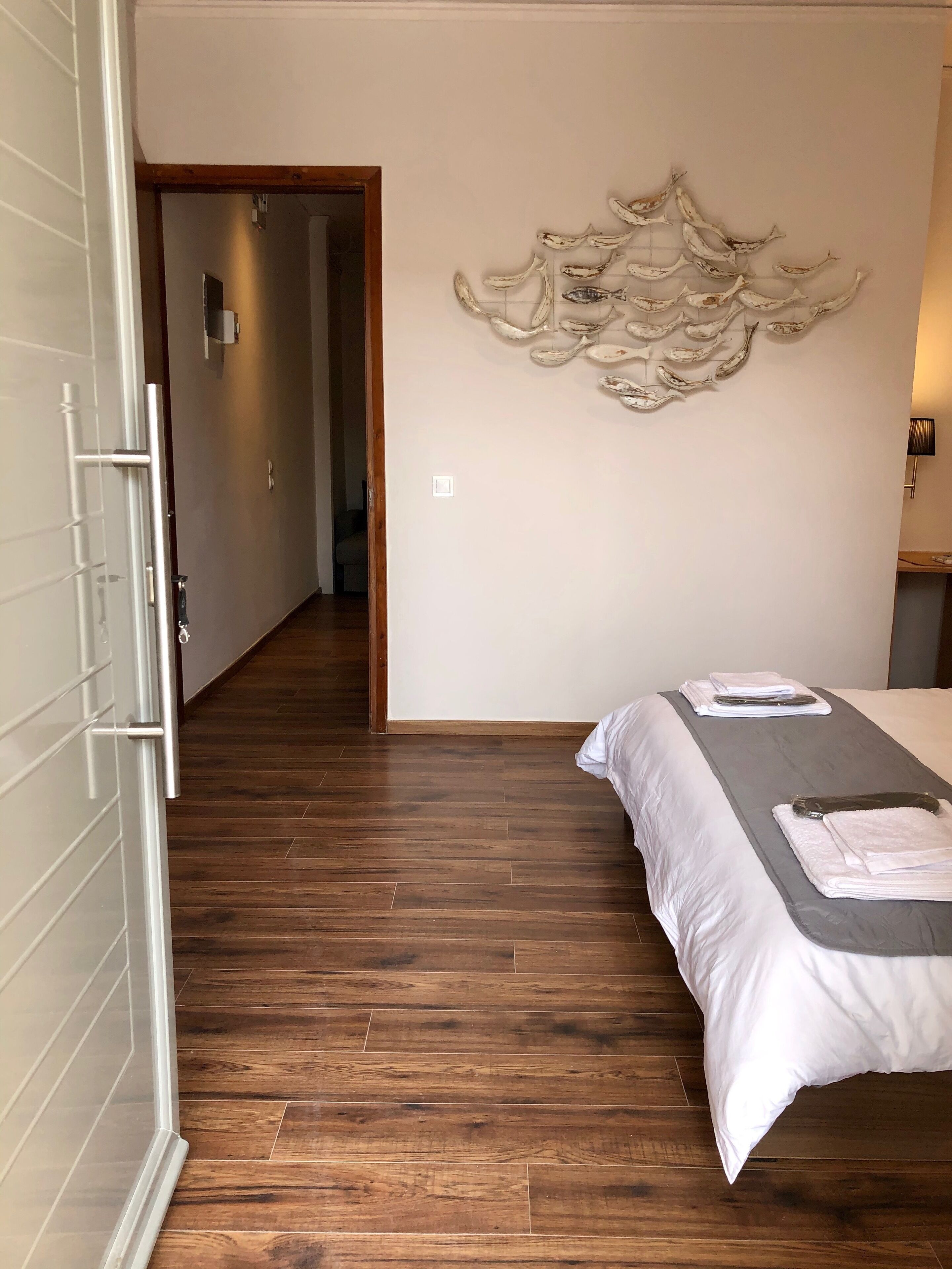 A hotel room with a wooden floor, a bed with white bedding, a towel rack, and a wall-mounted decorative piece.