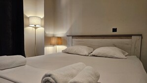 A neatly made bed with two rolled towels, a wooden headboard, and a wall-mounted lamp. - Lemon Tree Suites Near ATH Airport (Markopoulo Mesogaias)