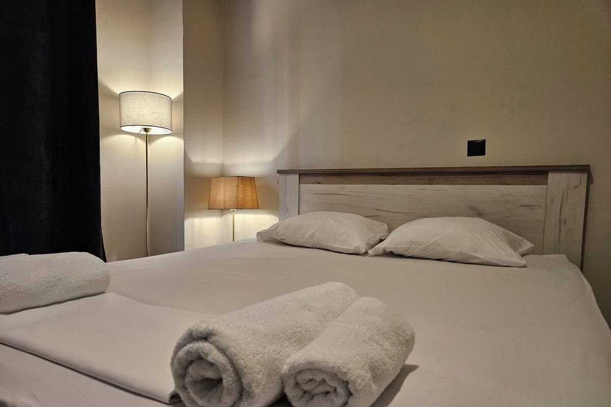 A neatly made bed with two rolled towels, a wooden headboard, and a wall-mounted lamp.
