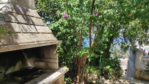 Un área de cocina al aire libre con una parrilla de piedra, rodeada de follaje y un pequeño árbol con flores rosadas.