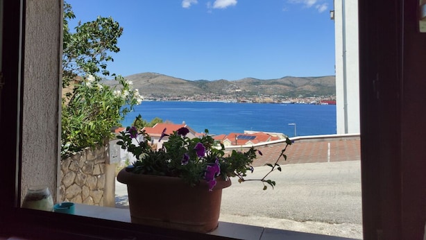 Vista costera desde una ventana con una planta en maceta, un muro de piedra y un cielo azul despejado.