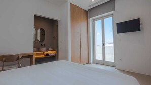 A modern bedroom with a large bed, a wooden desk, a mirror, a sink, and a TV mounted on the wall. - One Bedroom Suite, Amarelo Suites (Mykonos)