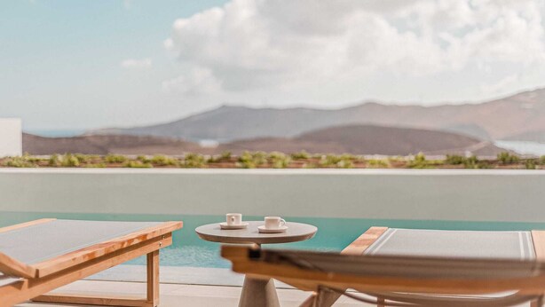 A poolside view with lounge chairs and a small table, overlooking a body of water and distant mountains. - One Bedroom Suite, Amarelo Suites (Mykonos)