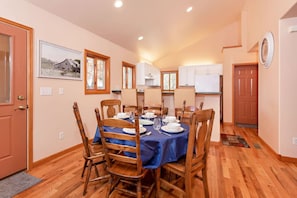 Un coin repas avec une table ronde dressée pour quatre personnes, des chaises en bois et une nappe bleue. On aperçoit, au fond, une cuisine avec des meubles blancs et un réfrigérateur.