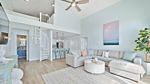 A spacious living area with a high ceiling, a staircase leading to an upper level, a kitchen with bar stools, and a large window with a view.