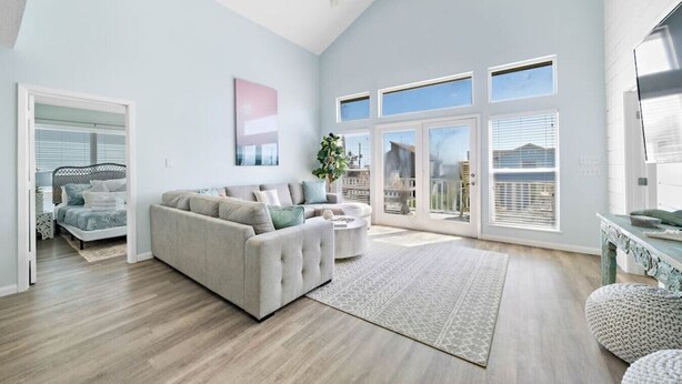A spacious living room with a large gray sectional sofa, a coffee table, and a ceiling fan. There are large windows and a sliding glass door leading to a balcony.