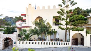 A white building with arched windows and a balcony, surrounded by greenery and potted plants. - Inviting Arokaria Apartment in Panormo (Mylopotamos)